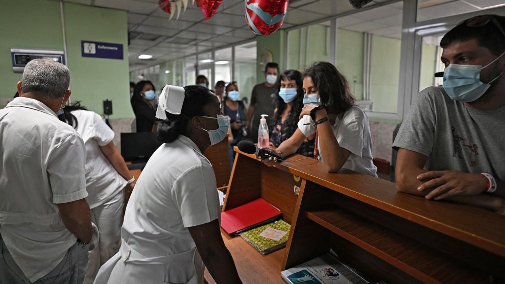 A person wearing a white medical uniform and nurse's cap is standing behind a wooden counter and speaking into a microphone held by another person. Several people wearing blue surgical masks stand on either side of the counter and in the background of the hallway. The following is written on the wall behind the counter: "nursing" Comes with medical symbol. Red and white heart-shaped balloons hang from the ceiling. The person on the right, wearing a gray T-shirt and blue surgical mask, leans against the counter and looks toward a person in medical attire.