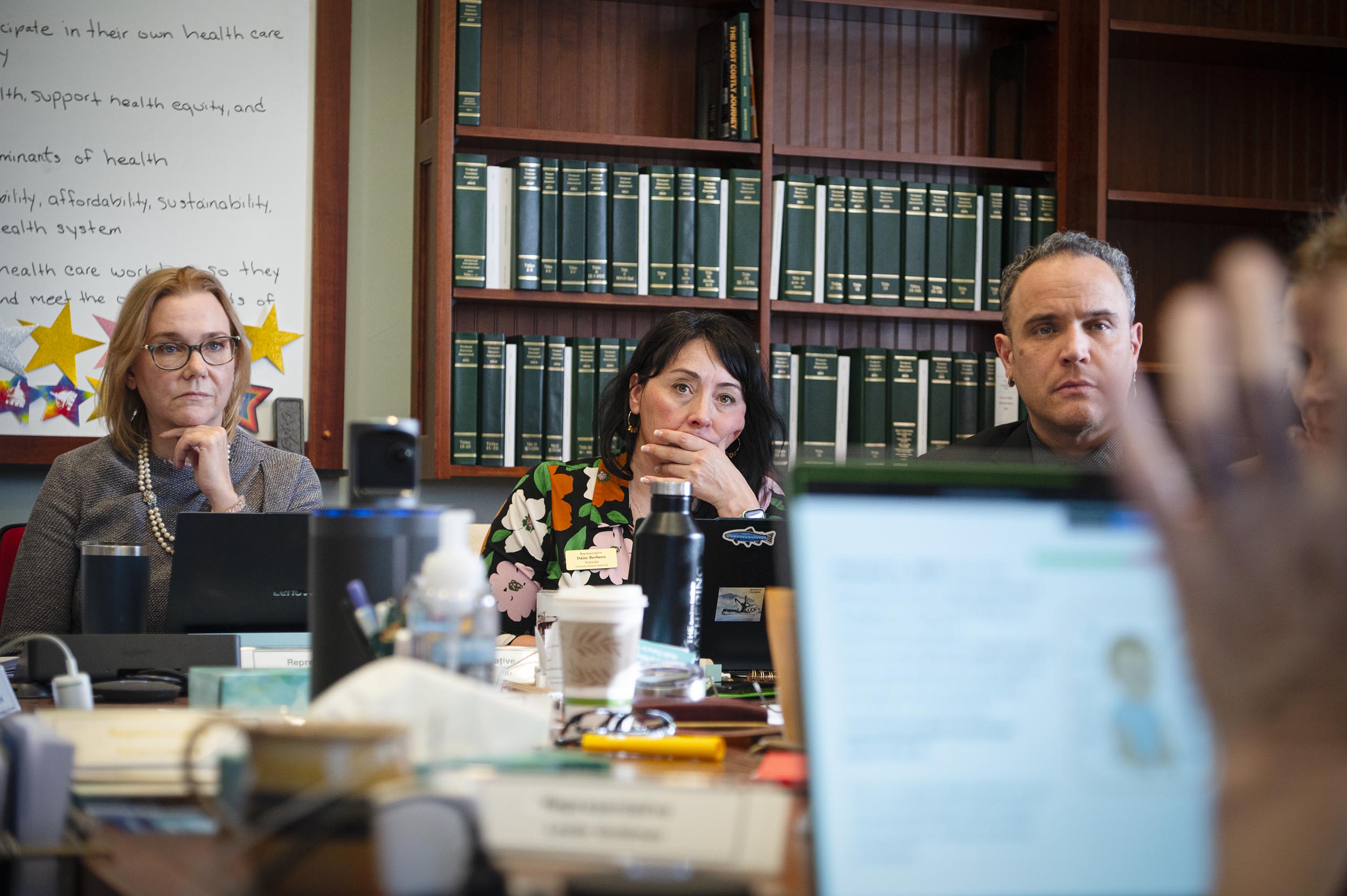 Three people are sitting at a conference table, listening intently during a meeting with bookshelves and a whiteboard in the background.